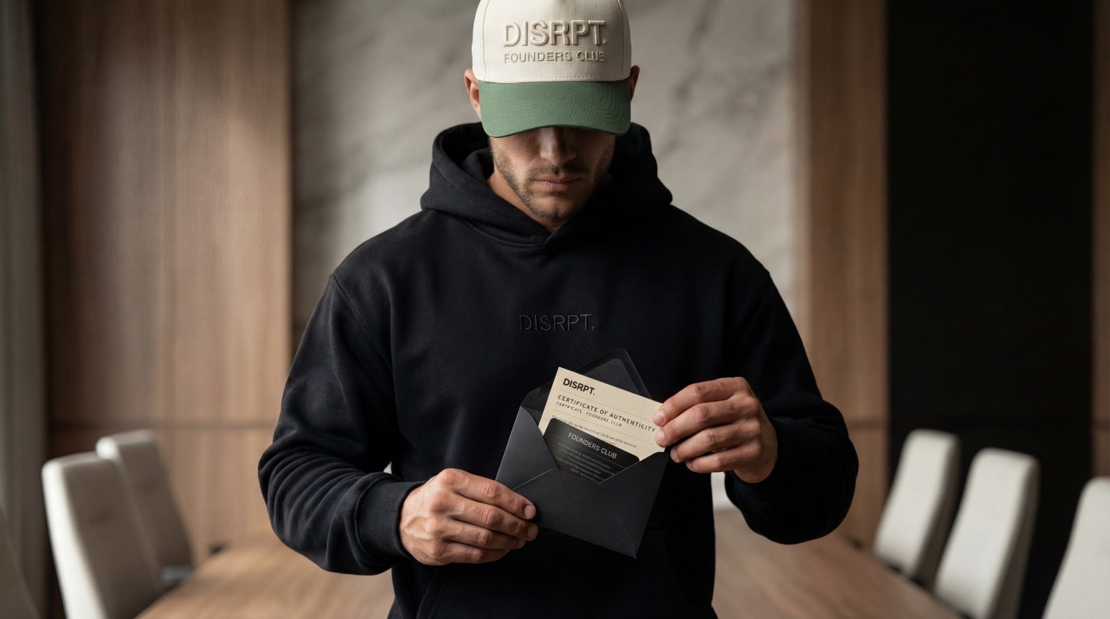 Man wearing a black DISRPT hoodie and mineral green Founders Club hat opening an envelope with a Founders Club card, representing exclusive access to a limited luxury streetwear drop.