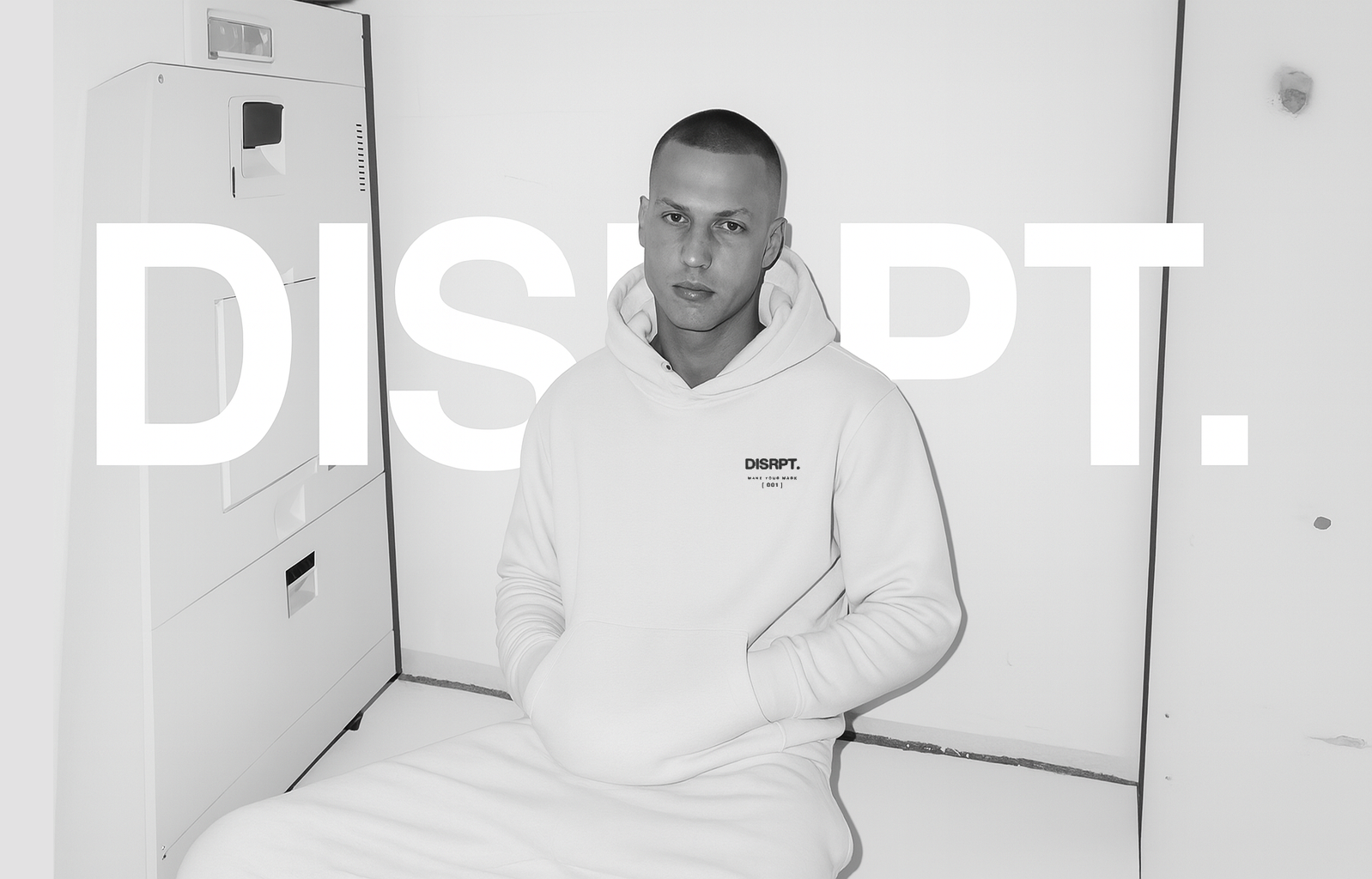 Person wearing white DISRPT hoodie sitting in minimalist studio setting with clean streetwear design